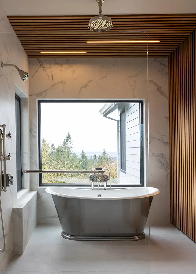 Primary bathroom remodel by JDI Construction with freestanding tub under a picture window, marble-look walls and wood slat accents
