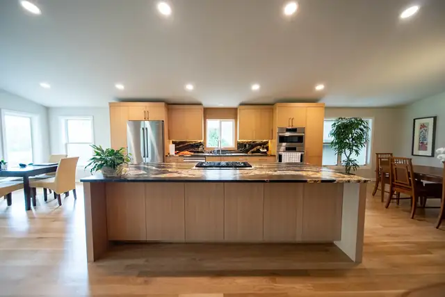 Open kitchen with a long wood-front island, a stone countertop, maple cabinets and a dining area by the windows