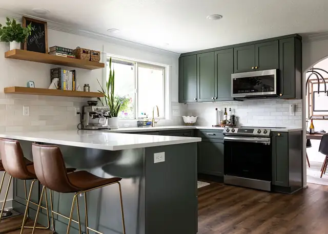 Kitchen with deep green shaker cabinets, a white quartz peninsula, leather bar stools and floating wood shelves
